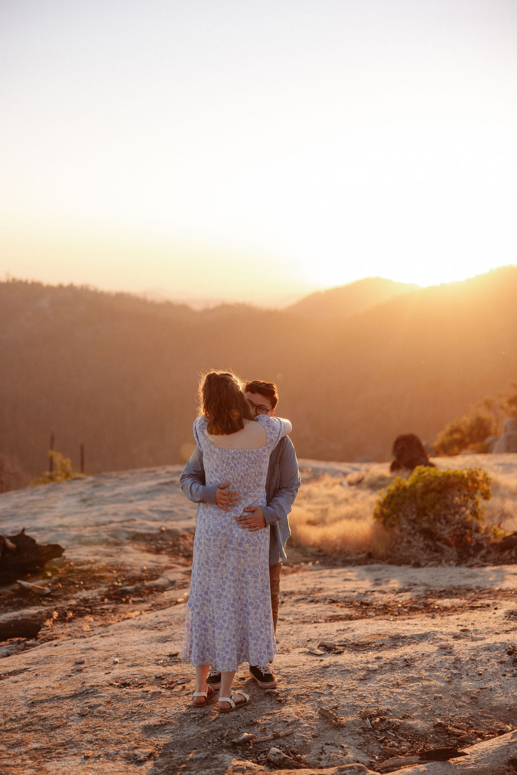 A couple embraces on a rocky landscape at sunset, with mountains and bright sunlight in the background.