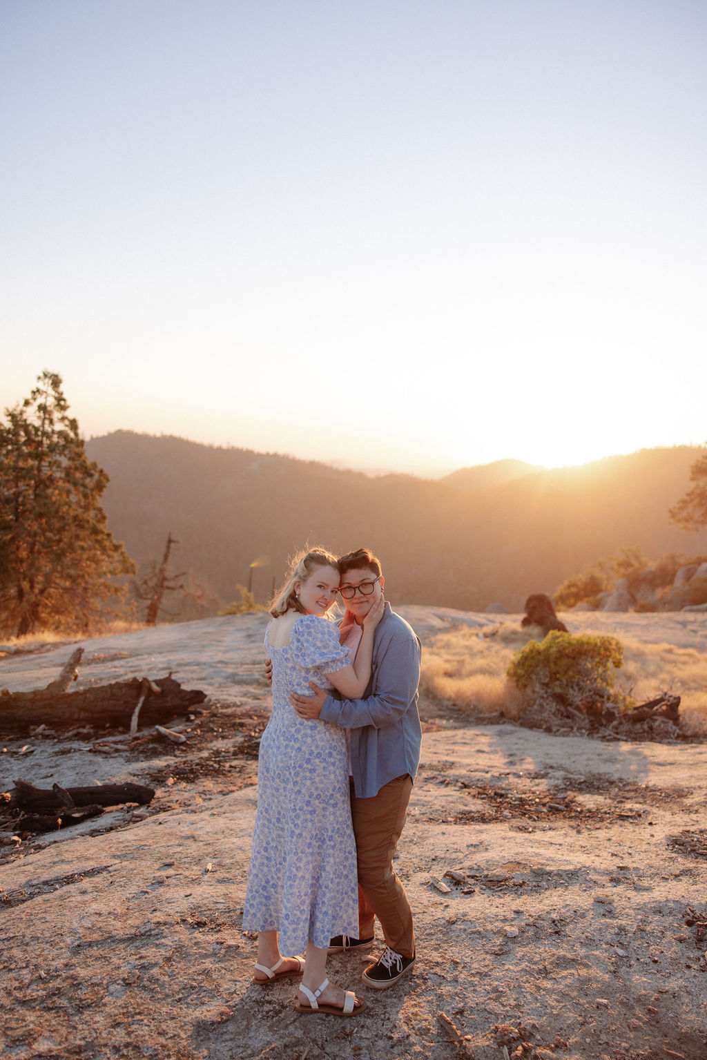 A couple embraces on a rocky landscape at sunset, with mountains and bright sunlight in the background.