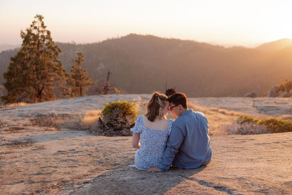 A couple sits close together on a rocky surface, facing a scenic mountain landscape at sunset.