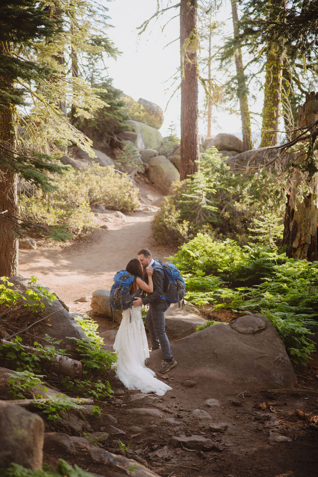 Two people with backpacks walk on a rocky forest trail surrounded by tall trees.