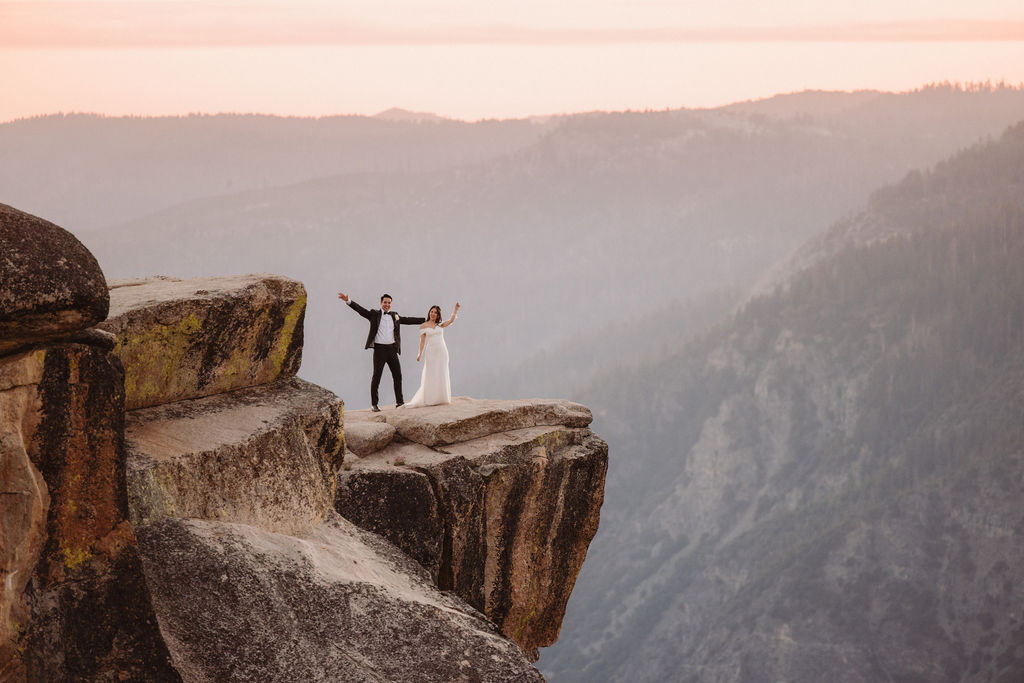 A couple in formal attire stands on the edge of a rocky cliff at sunset, with mountains and a hazy sky in the background for their unique elopement