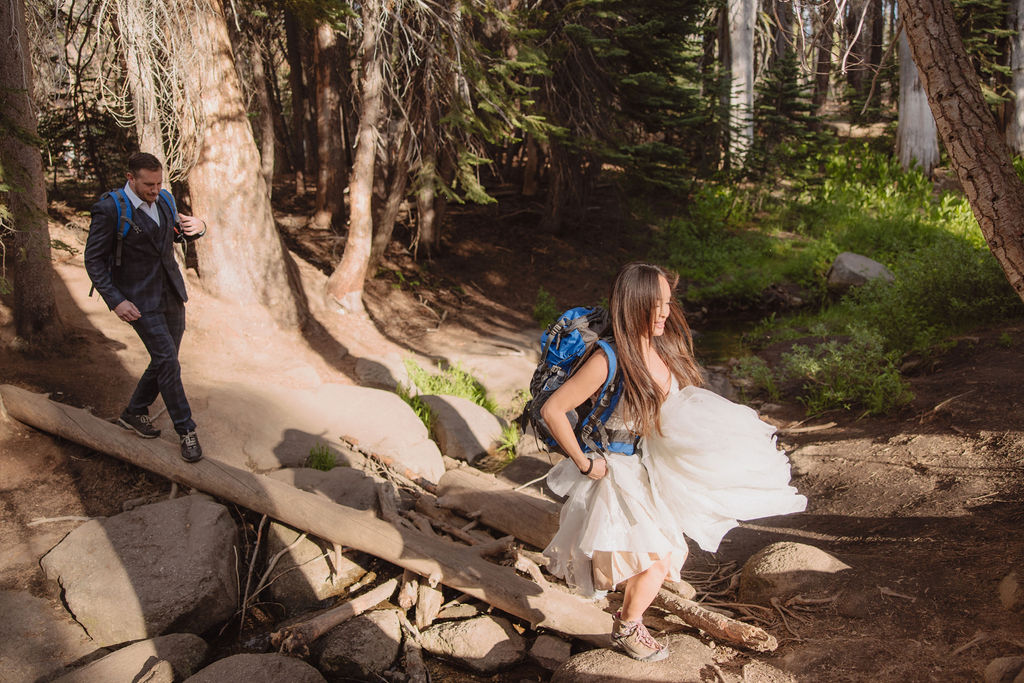 Two people with backpacks walk on a rocky forest trail surrounded by tall trees.