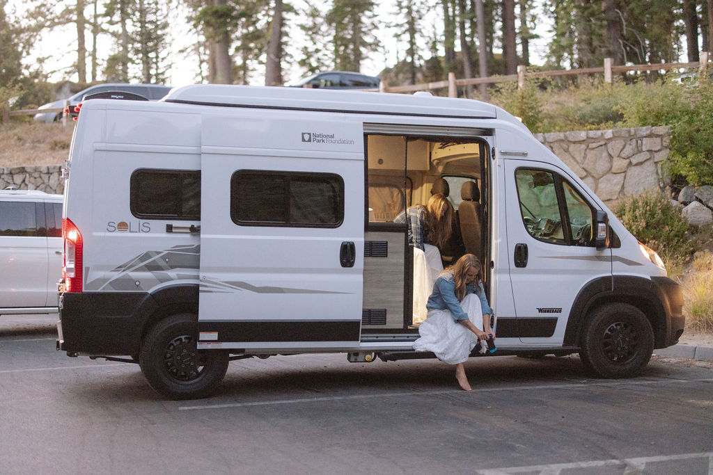 A woman sits on the step of a white camper van with its side door open, parked in a lot with trees and rocks in the background.