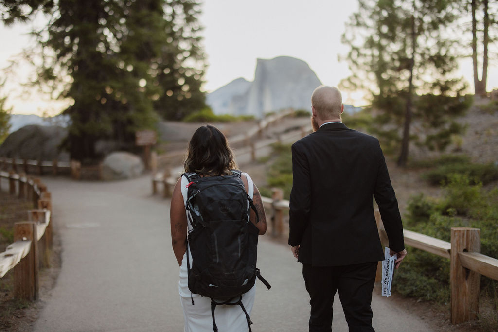 Two people walk on a paved trail in a natural setting; one wears a backpack and the other is in a suit holding a book or folder. Trees and mountains are in the background.