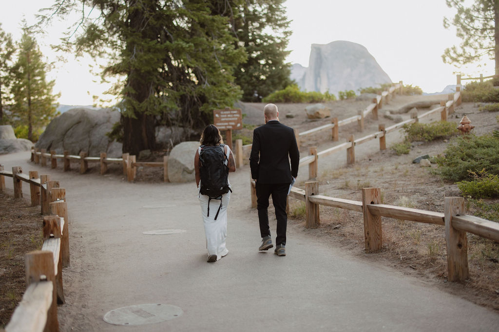 Two people walk on a paved trail in a natural setting; one wears a backpack and the other is in a suit holding a book or folder. Trees and mountains are in the background.