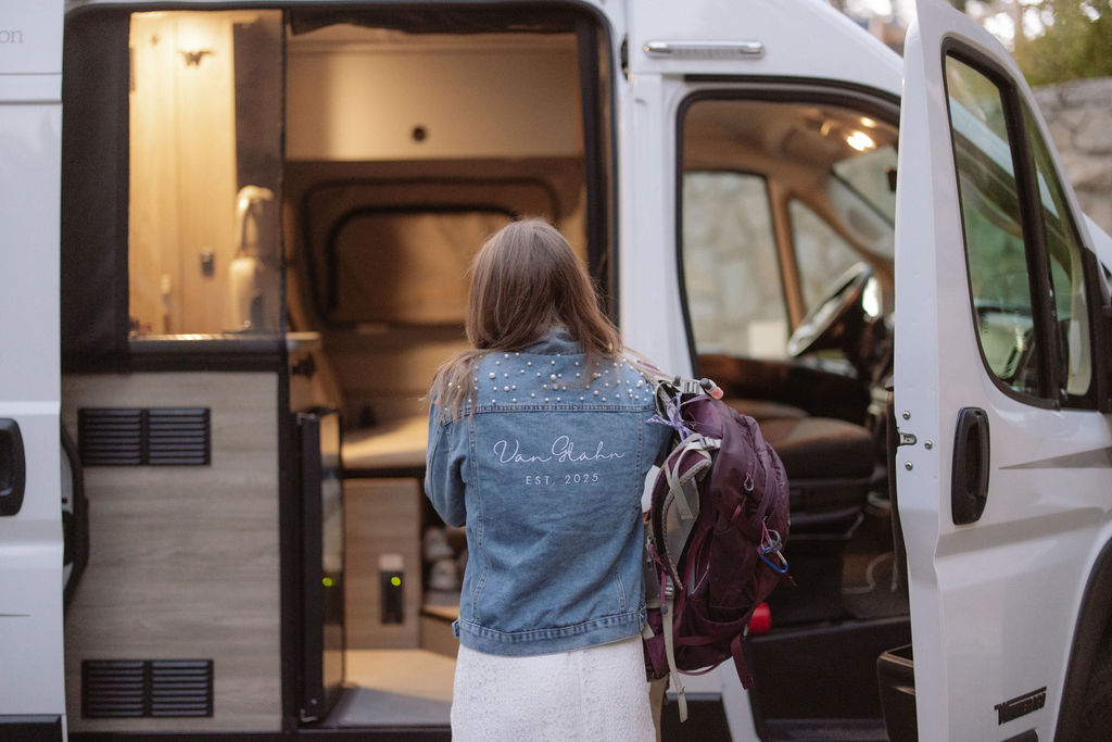 A woman sits on the step of a white camper van with its side door open, parked in a lot with trees and rocks in the background.