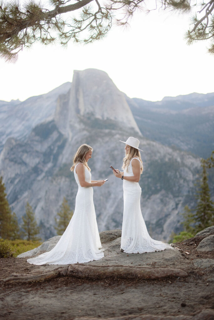 Two women in white dresses stand on a rocky ledge with a mountain landscape in the background; one is wearing a hat and looking at a phone for a Yosemite elopement