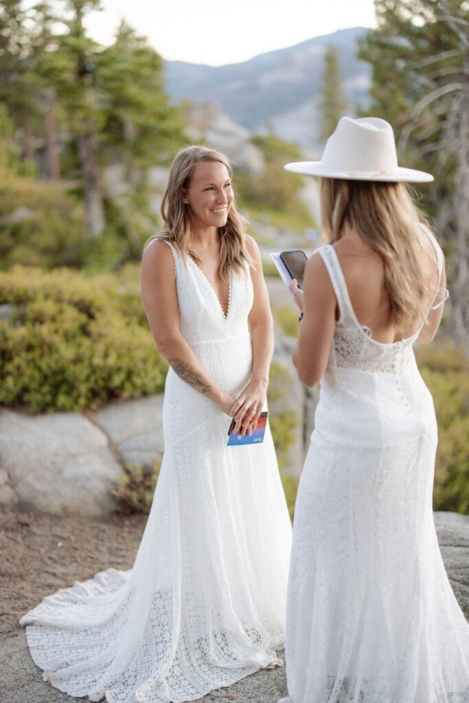 Two women in white dresses stand on a rocky ledge with a mountain landscape in the background; one is wearing a hat and looking at a phone for a Yosemite elopement