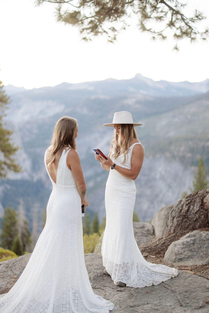 Two women in white dresses stand on a rocky ledge with a mountain landscape in the background; one is wearing a hat and looking at a phone for a Yosemite elopement