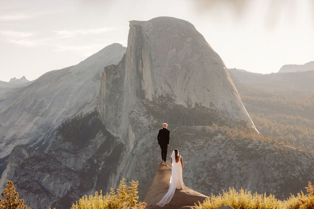 A couple in wedding attire stands on a rocky ledge with Half Dome and mountain scenery in the background for a glacier point elopement