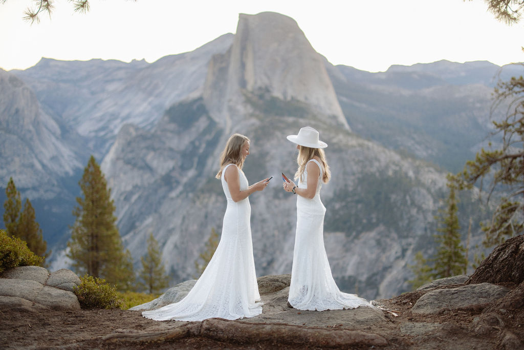 Two women in white dresses stand on a rocky ledge with a mountain landscape in the background; one is wearing a hat and looking at a phone for a Yosemite elopement