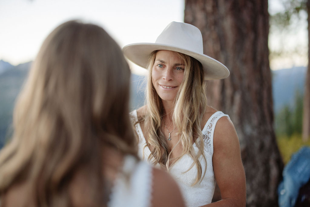 Two women in white dresses stand on a rocky ledge with a mountain landscape in the background; one is wearing a hat and looking at a phone for a Yosemite elopement