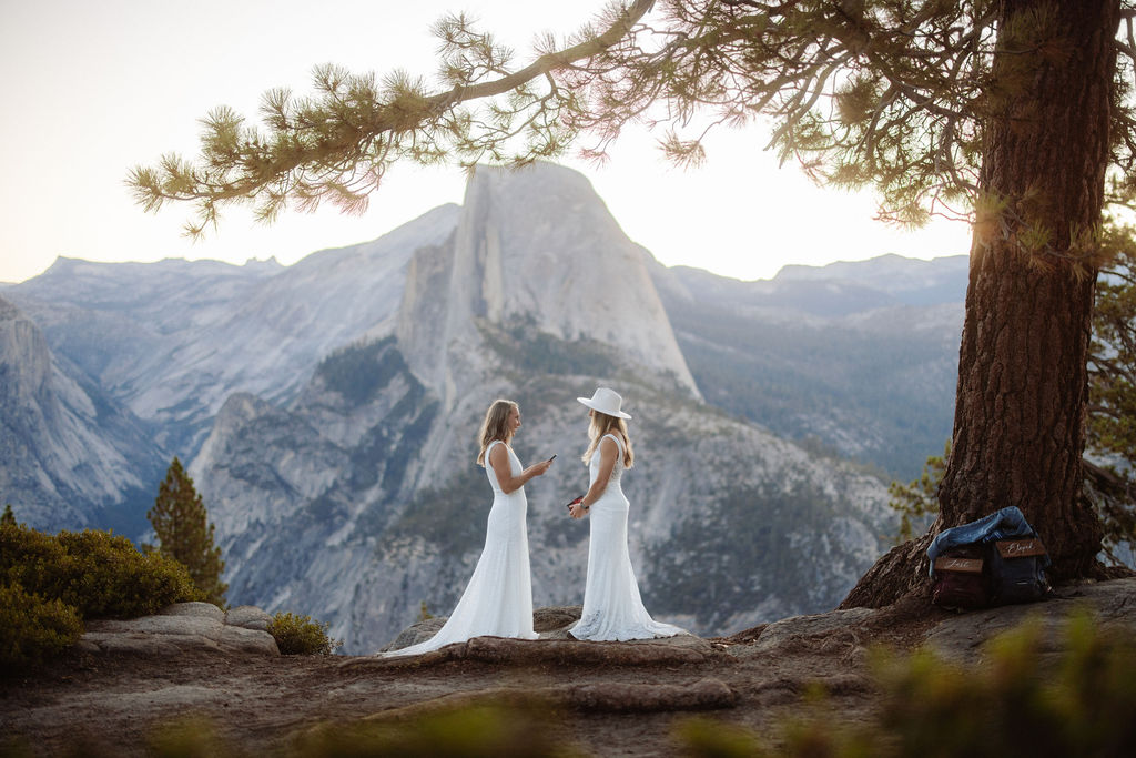 Two women in white dresses stand on a rocky ledge with a mountain landscape in the background; one is wearing a hat and looking at a phone for a Yosemite elopement