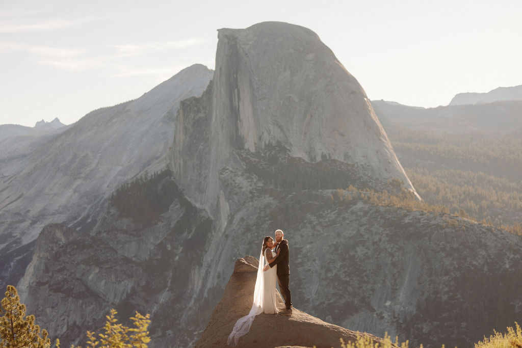 A couple in wedding attire stands on a rocky ledge with Half Dome and mountain scenery in the background for a glacier point elopement