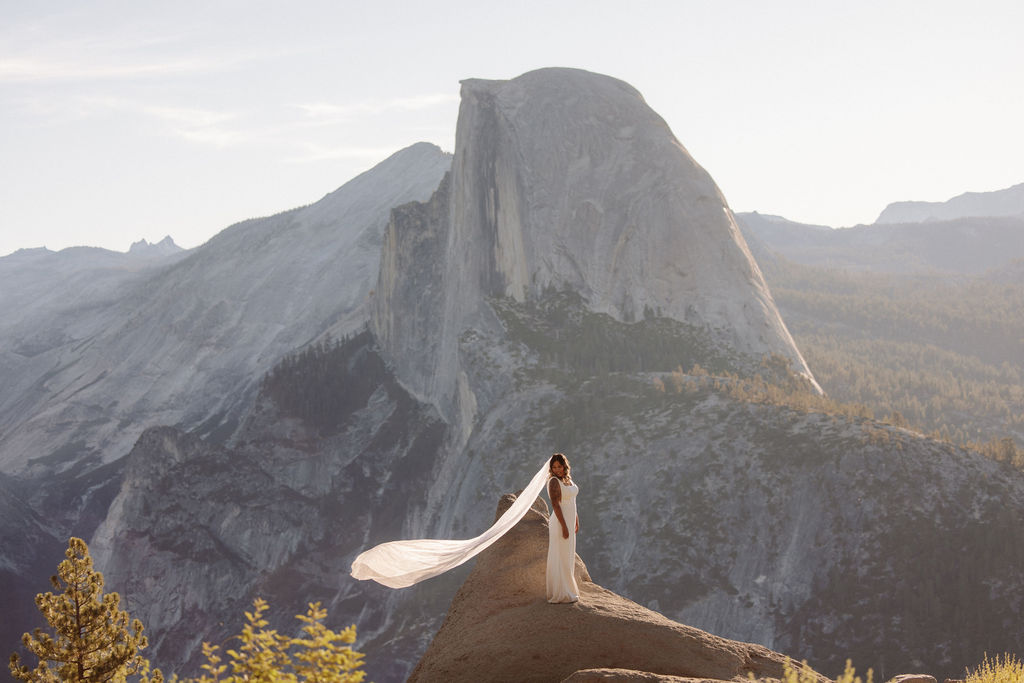 A couple in wedding attire stands on a rocky ledge with Half Dome and mountain scenery in the background for a glacier point elopement
