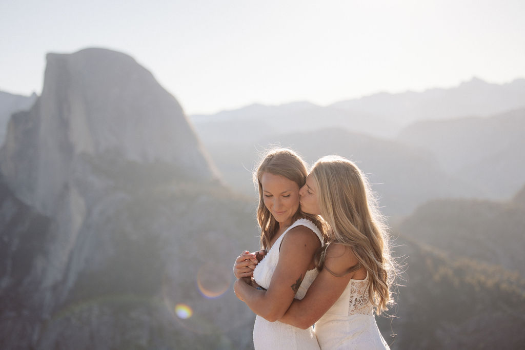 Two women in white dresses embrace and share a kiss on the cheek with a mountainous landscape and sunlight in the background.