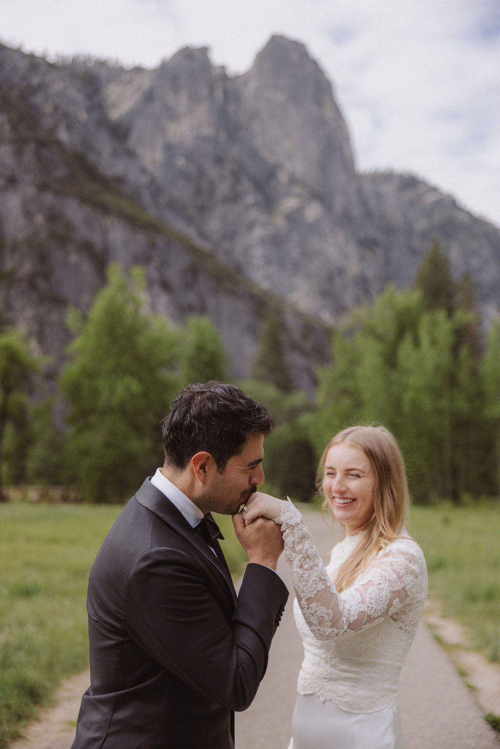 A bride and groom stand together in a grassy field with pine trees and a large mountain in the background, both facing each other and holding hands for their elopement in Yosemite | elopement budget