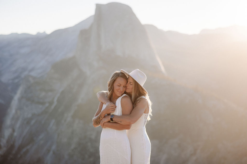 Two women in white dresses embrace and share a kiss on the cheek with a mountainous landscape and sunlight in the background.