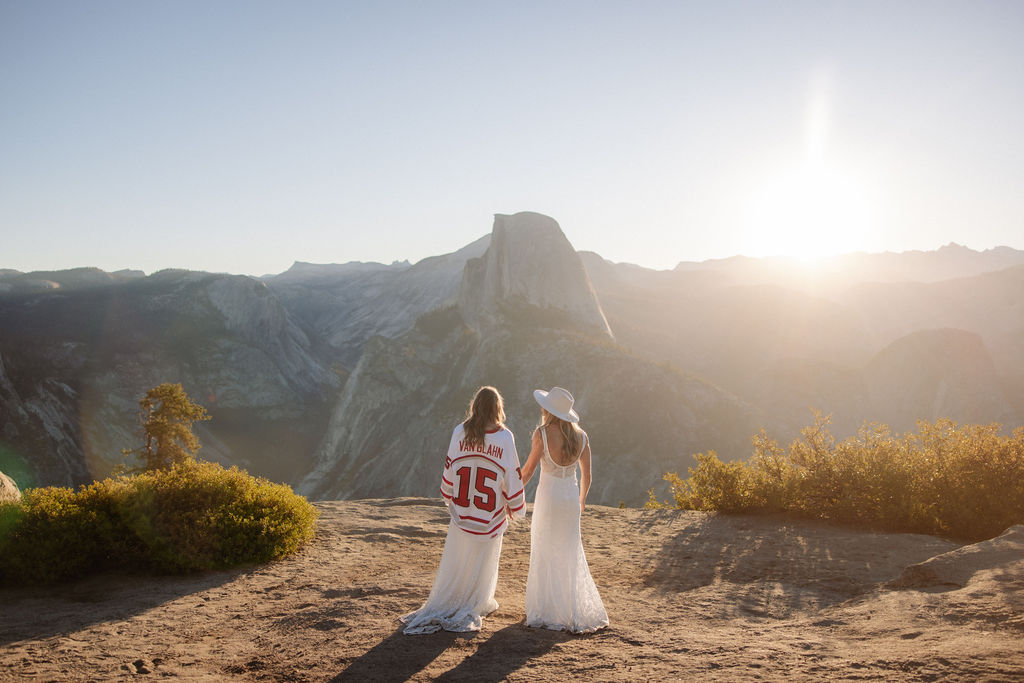 Two women in white dresses stand on a mountain overlook at sunrise or sunset; one wears a hat and the other a sports jersey with the number 15.