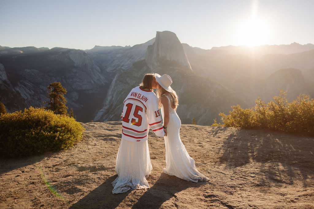 Two women in white dresses stand on a mountain overlook at sunrise or sunset; one wears a hat and the other a sports jersey with the number 15.