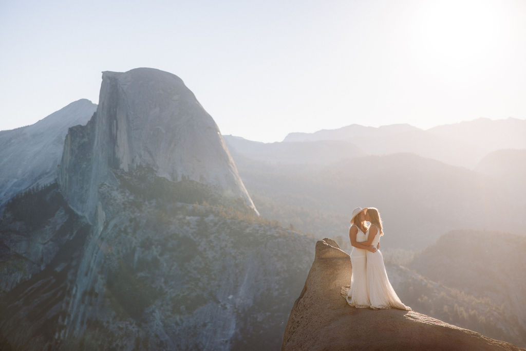 Two people in wedding attire stand holding hands on the edge of a rocky cliff with mountains visible in the background for a Yosemite elopement