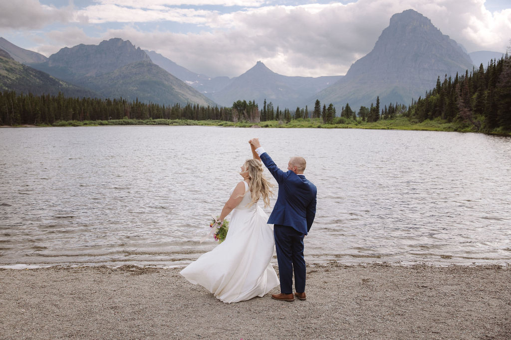 A bride and groom dance on a lakeshore with mountains and trees in the background under a cloudy sky.