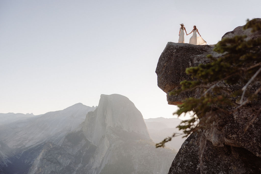 Two people in wedding attire stand holding hands on the edge of a rocky cliff with mountains visible in the background for a Yosemite elopement