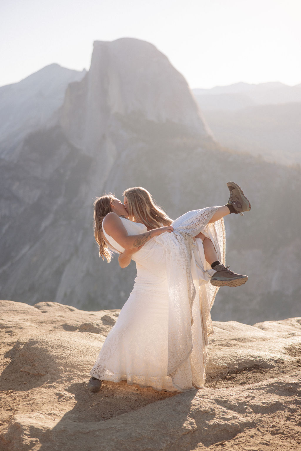 Two people in wedding attire stand holding hands on the edge of a rocky cliff with mountains visible in the background for a Yosemite elopement