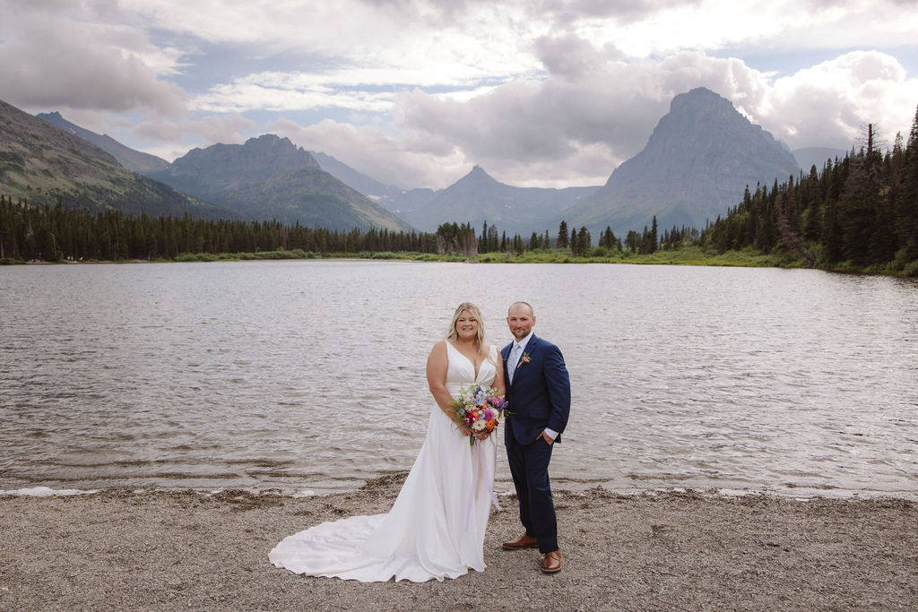 A bride and groom stand together by a lake, with mountains and trees in the background under a cloudy sky for a wedding at Glacier national park