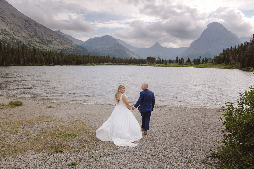 A bride and groom stand together by a lake, with mountains and trees in the background under a cloudy sky for a wedding at Glacier national park