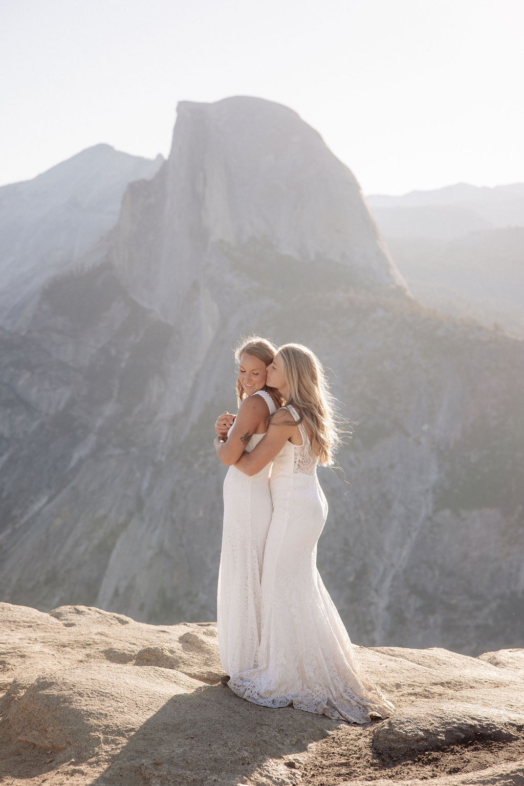 Two people in wedding attire stand holding hands on the edge of a rocky cliff with mountains visible in the background for a Yosemite elopement