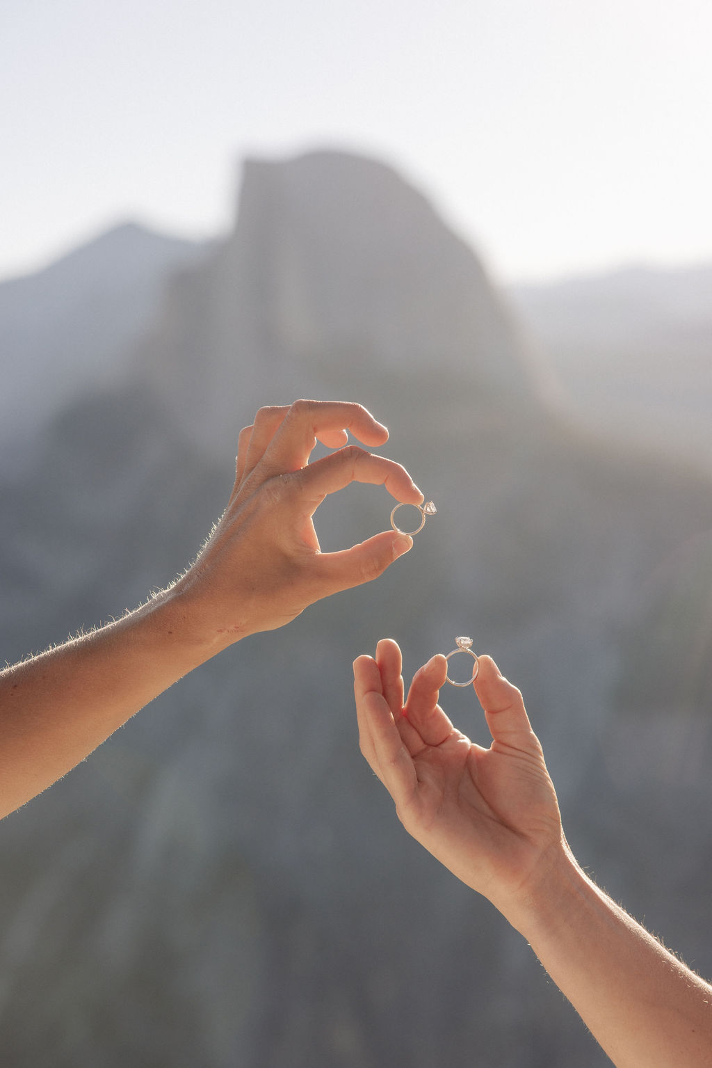 Two people in wedding attire stand holding hands on the edge of a rocky cliff with mountains visible in the background for a Yosemite elopement