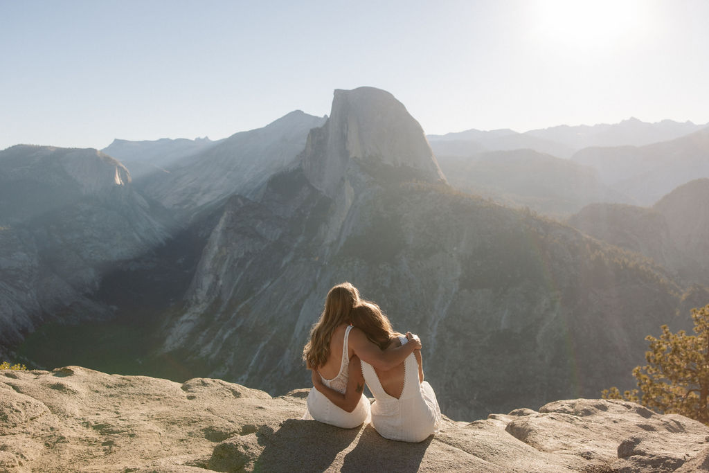 Two people in wedding attire stand holding hands on the edge of a rocky cliff with mountains visible in the background for a Yosemite elopement