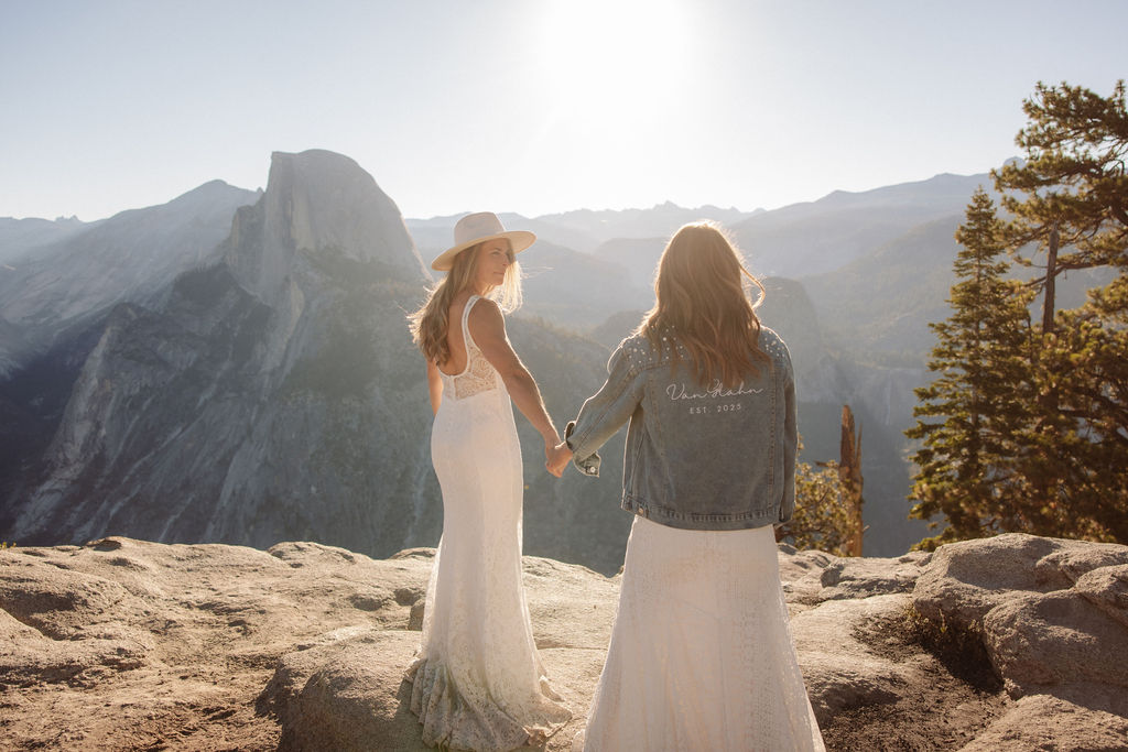Two people in wedding attire stand holding hands on the edge of a rocky cliff with mountains visible in the background for a Yosemite elopement