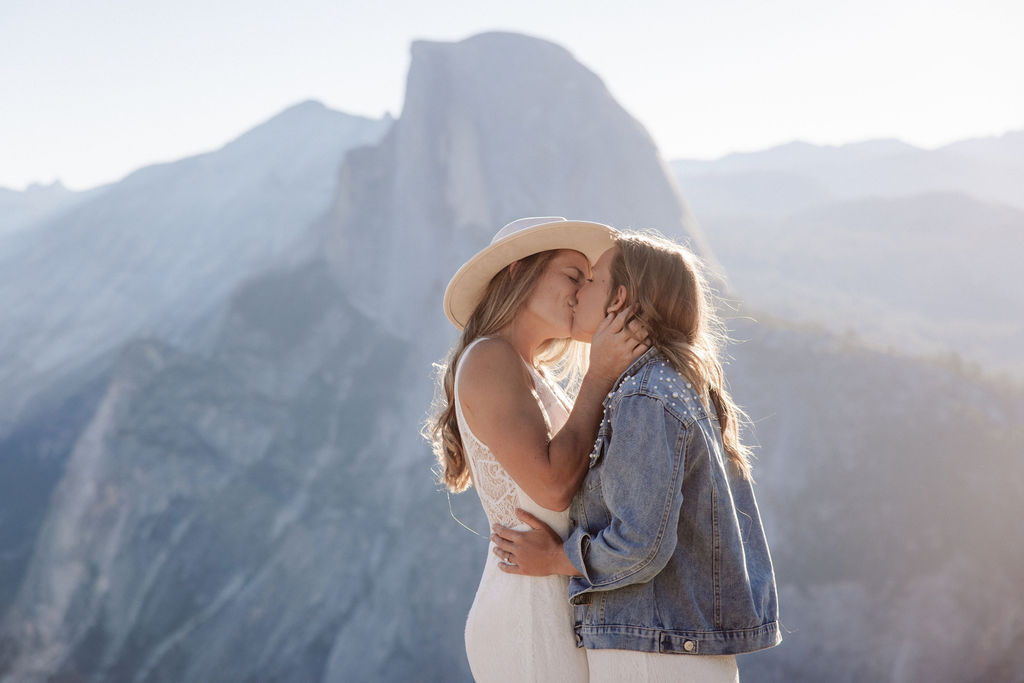 Two people in wedding attire stand holding hands on the edge of a rocky cliff with mountains visible in the background for a Yosemite elopement