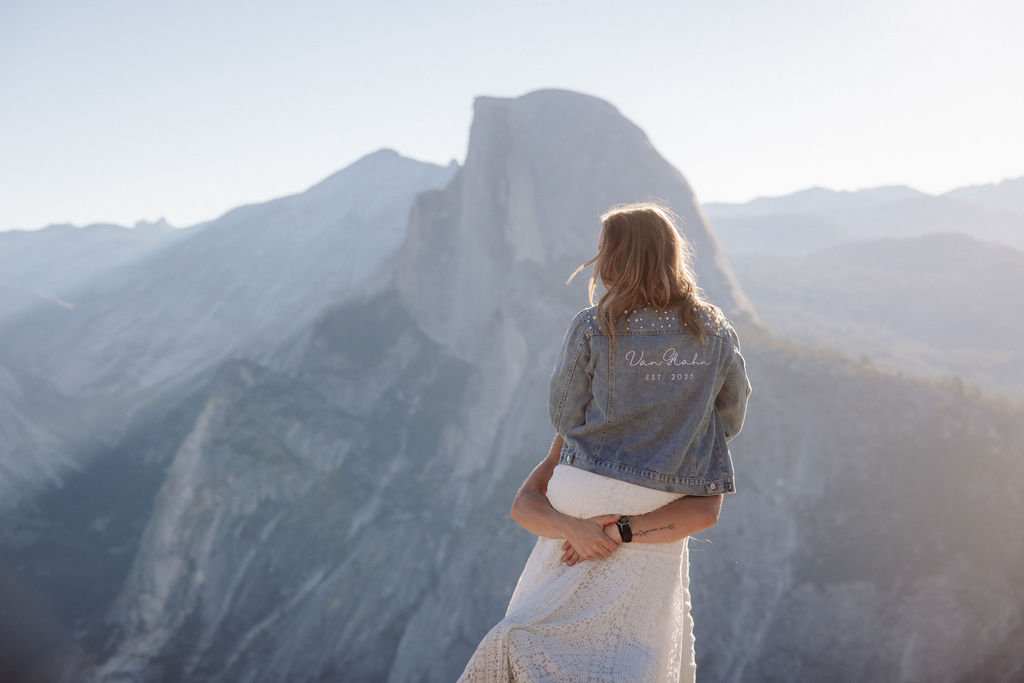 Two people in wedding attire stand holding hands on the edge of a rocky cliff with mountains visible in the background for a Yosemite elopement