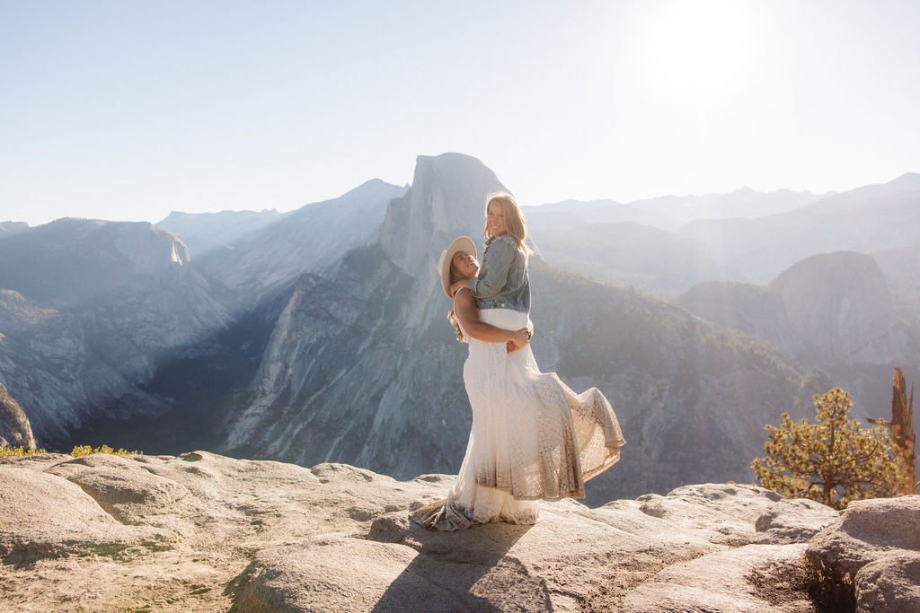Two people in wedding attire stand holding hands on the edge of a rocky cliff with mountains visible in the background for a Yosemite elopement