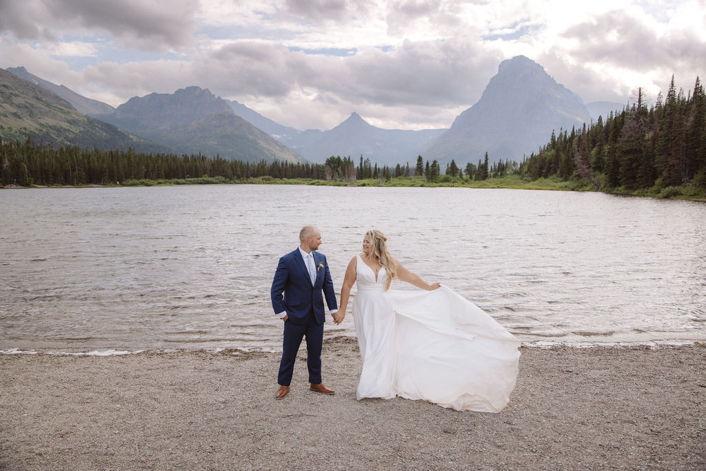 A bride and groom stand together by a lake, with mountains and trees in the background under a cloudy sky for a wedding at Glacier national park