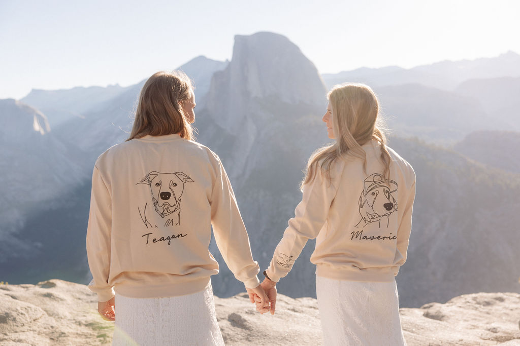 Two women stand outdoors holding hands, both wearing light jackets with dog illustrations and names on the back, with mountains visible in the background.