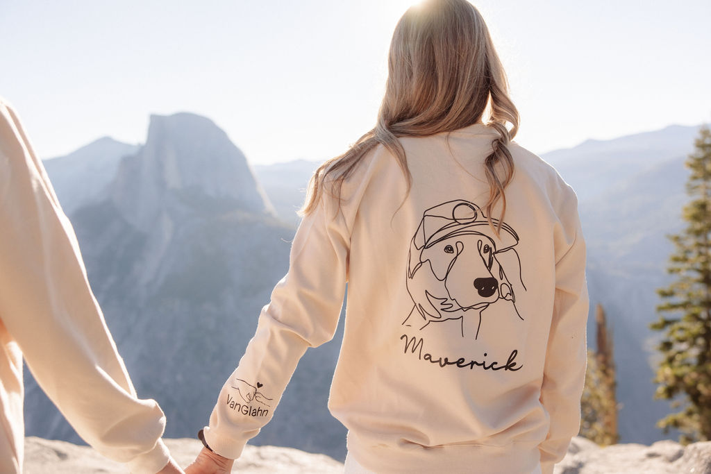 Two women stand outdoors holding hands, both wearing light jackets with dog illustrations and names on the back, with mountains visible in the background.