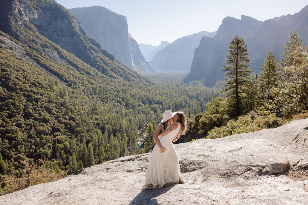 A couple in wedding attire shares a kiss on a rocky overlook, with a forested valley and mountains in the background for their Yosemite elopement