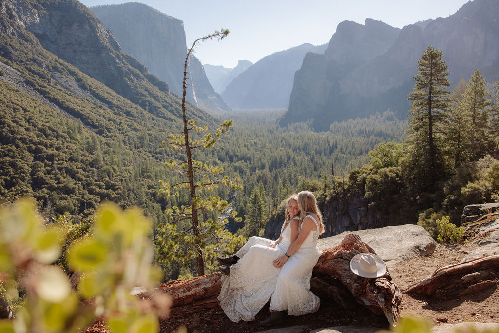 A couple in wedding attire shares a kiss on a rocky overlook, with a forested valley and mountains in the background for their Yosemite elopement