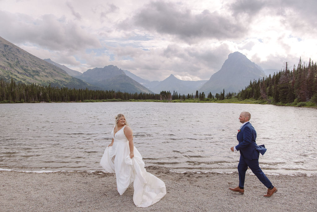 A bride and groom stand together by a lake, with mountains and trees in the background under a cloudy sky for a wedding at Glacier national park