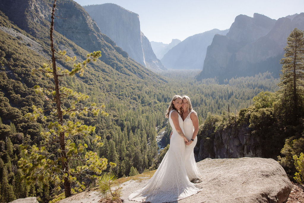A couple in wedding attire shares a kiss on a rocky overlook, with a forested valley and mountains in the background for their Yosemite elopement
