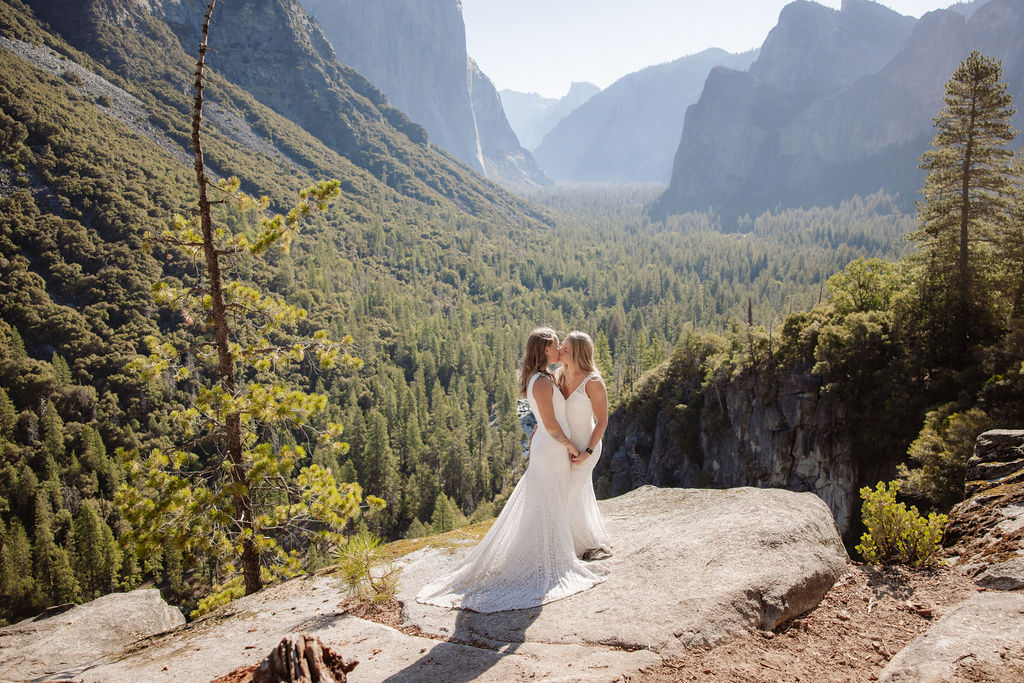 A couple in wedding attire shares a kiss on a rocky overlook, with a forested valley and mountains in the background for their Yosemite elopement