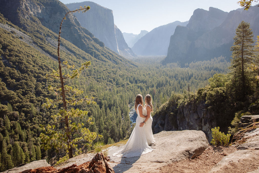 A couple in wedding attire shares a kiss on a rocky overlook, with a forested valley and mountains in the background for their Yosemite elopement