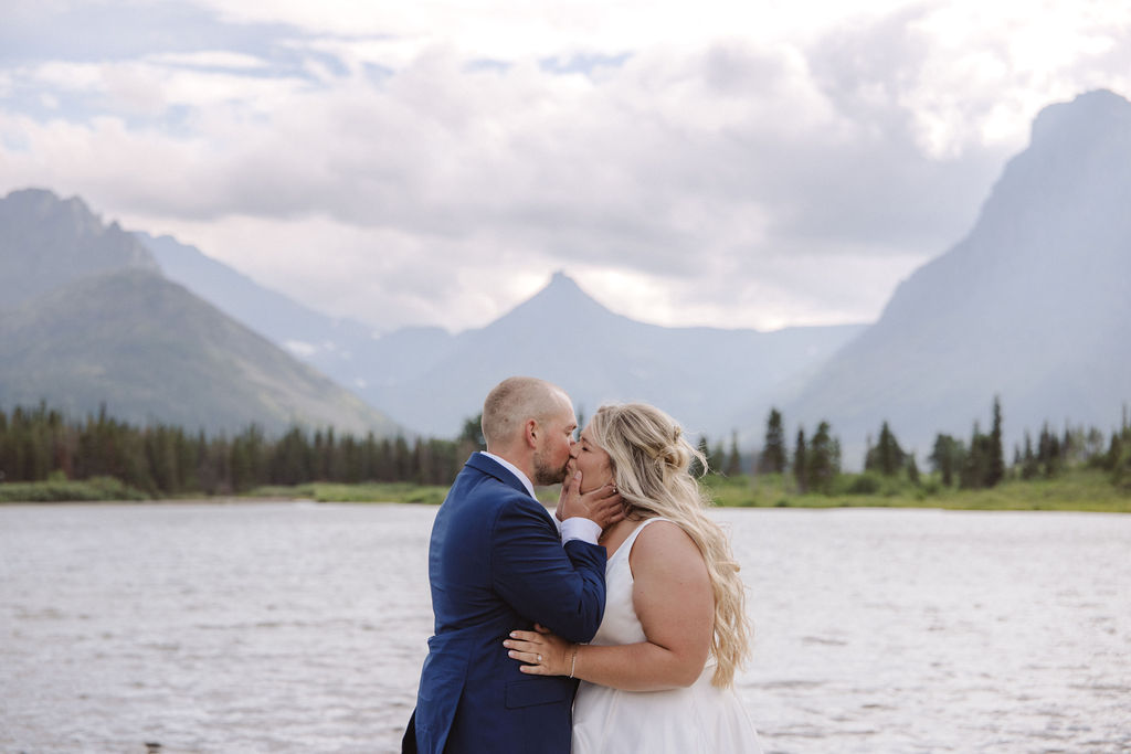 A bride and groom stand together by a lake, with mountains and trees in the background under a cloudy sky for a wedding at Glacier national park