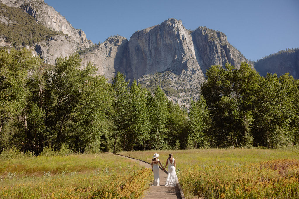 Two people holding hands walk on a wooden path through a grassy field, with tall trees and rocky mountains in the background under a clear blue sky.