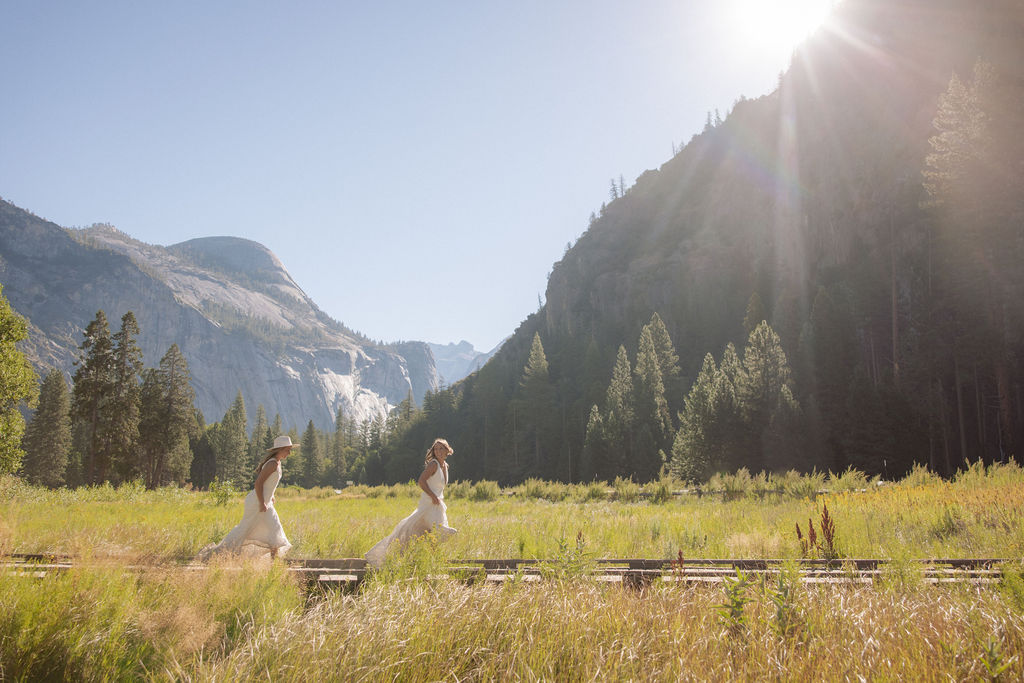 Two people holding hands walk on a wooden path through a grassy field, with tall trees and rocky mountains in the background under a clear blue sky.