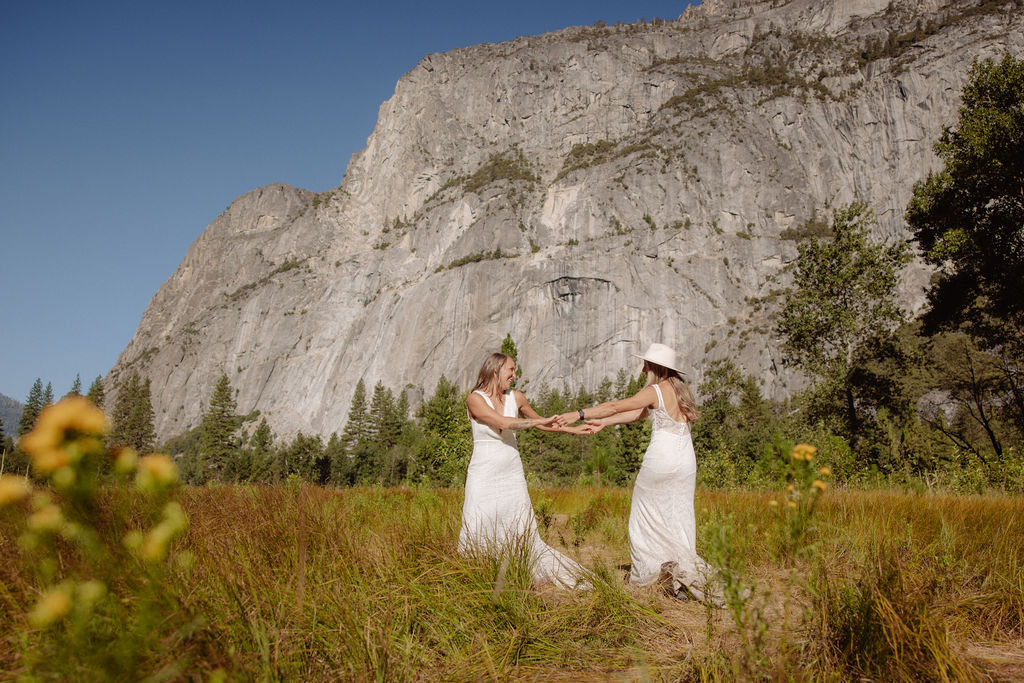 Two people holding hands walk on a wooden path through a grassy field, with tall trees and rocky mountains in the background under a clear blue sky.